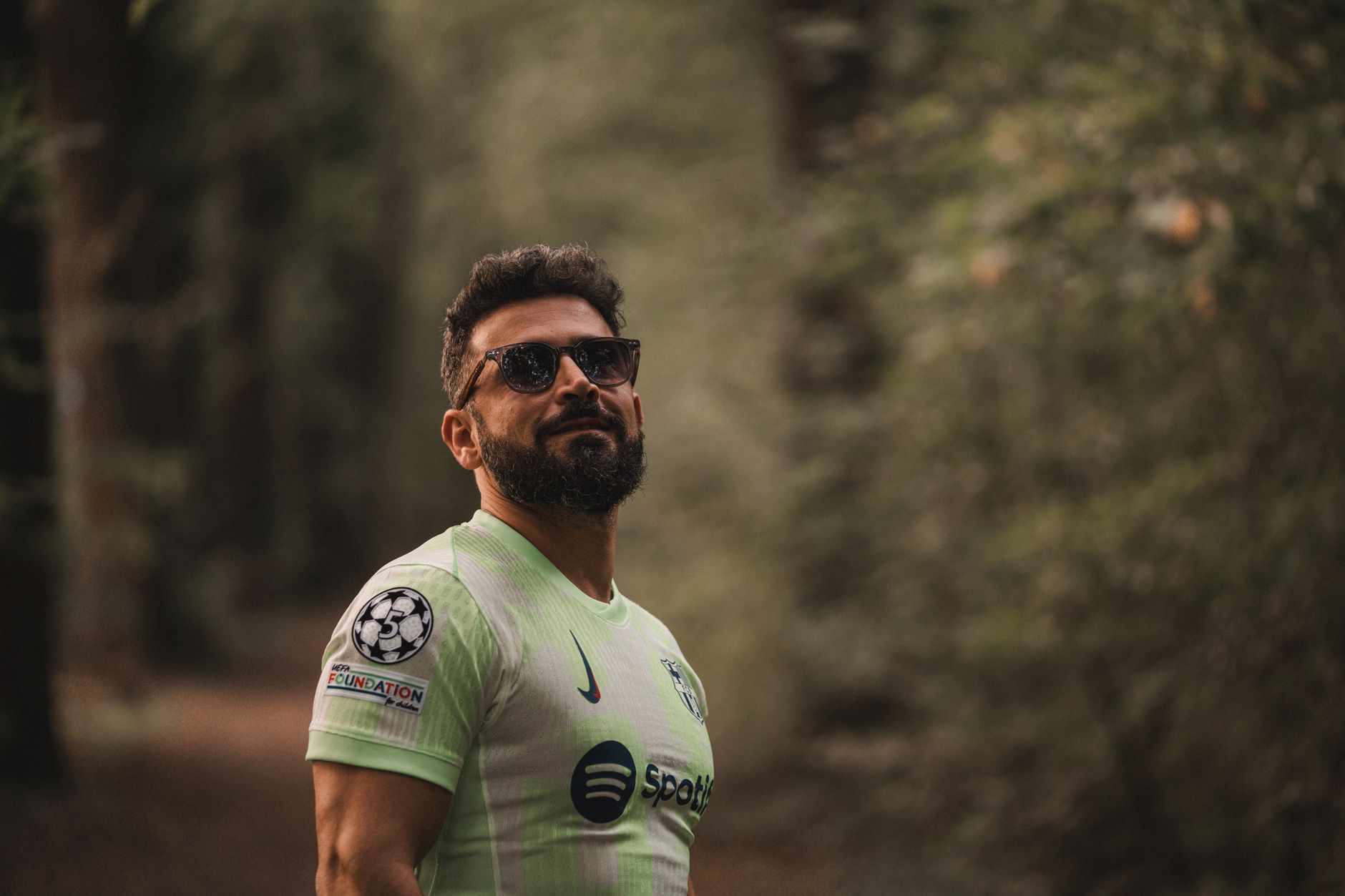 Seorang pria berjanggut dalam jersey sepak bola berdiri dengan percaya diri di lingkungan hutan yang tenang.