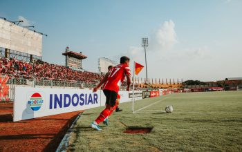 Seorang pemain sepak bola melakukan tendangan sudut di stadion Indonesia yang ramai dan dipenuhi penggemar.