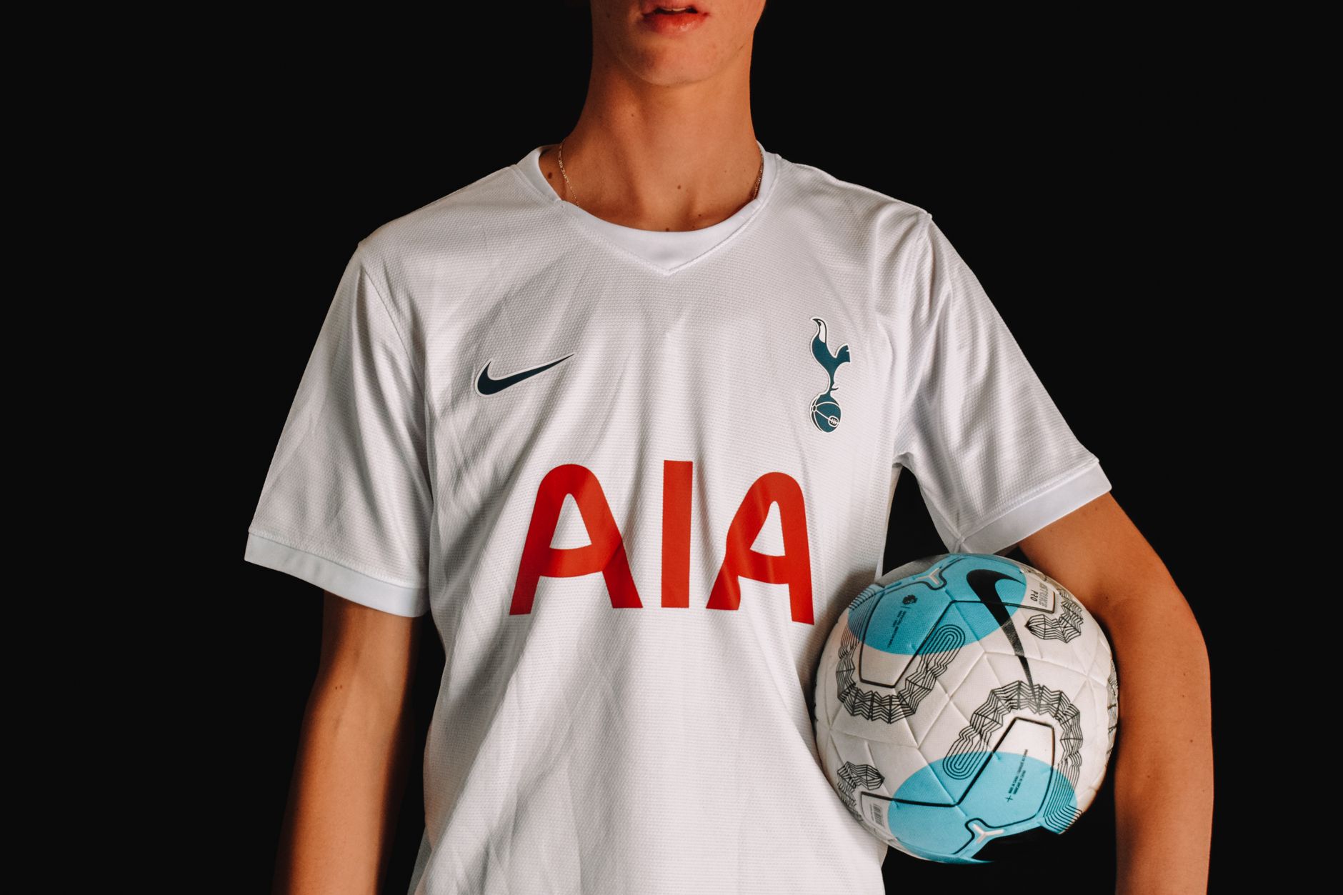Pemain sepak bola dewasa dengan jersey Tottenham Hotspur memegang bola di latar belakang gelap.