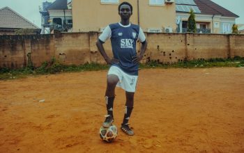 Seorang pemain sepak bola remaja dalam seragam berpose dengan bola di lapangan tanah.