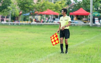 Seorang hakim garis sepak bola berseragam berdiri di lapangan berumput dengan bendera selama pertandingan siang hari.