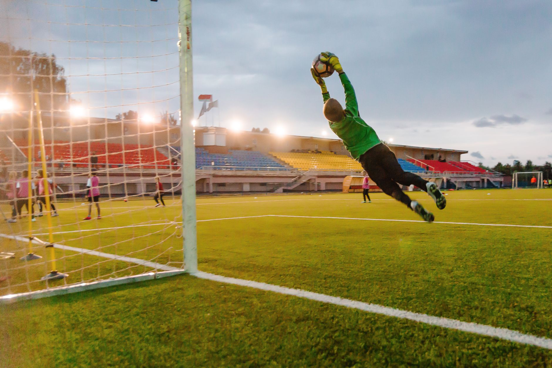 Kiper melakukan penyelamatan menyelam di lapangan sepak bola selama pertandingan saat matahari terbenam.