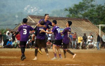 Pemain sepak bola merayakan gol dalam pertandingan luar ruangan yang berdebu dengan penonton.