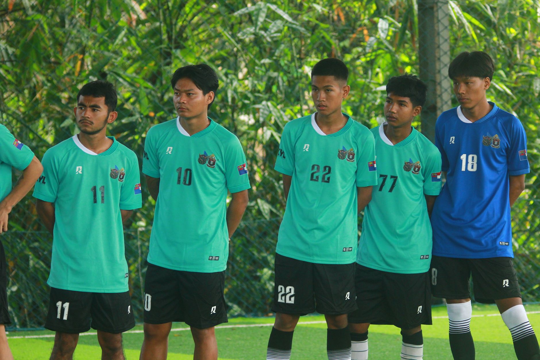 Sekelompok pemain sepak bola muda laki-laki mengenakan jersey hijau berdiri di luar ruangan di Myanmar.