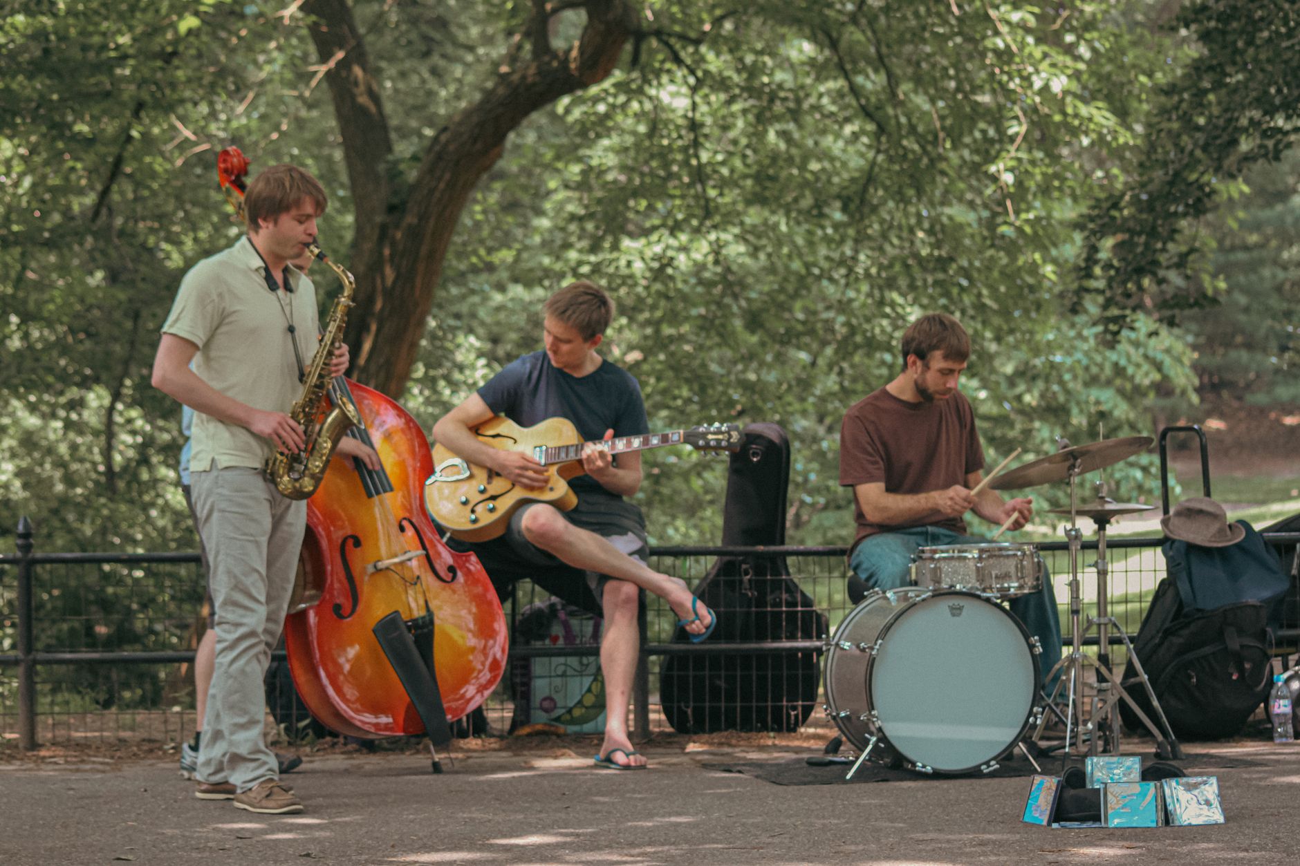 Tiga musisi tampil di taman Kota New York, memainkan saksofon, gitar, dan drum.
