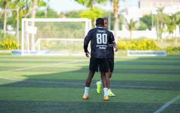 Dua pria berpartisipasi dalam sesi latihan sepak bola di luar ruangan pada hari yang cerah, berlatih keterampilan bermain bola.