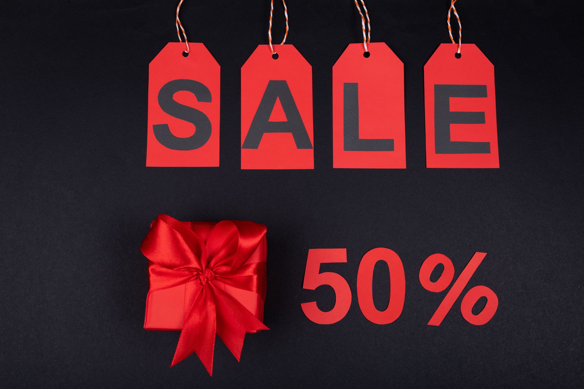 Kotak hadiah merah di samping label penjualan dan tanda 50% di latar belakang hitam, ideal untuk promosi.