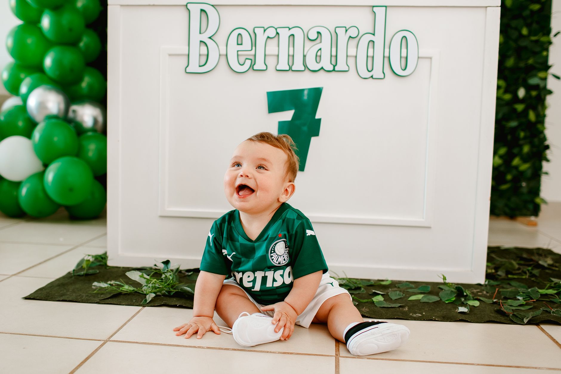 Bayi yang imut dan tersenyum mengenakan jersey sepak bola hijau, duduk di dalam ruangan dengan balon.