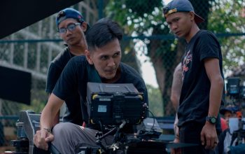 Sekelompok pembuat film yang meninjau rekaman di lokasi syuting luar ruangan, fokus pada perlengkapan kamera.