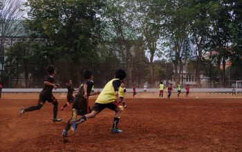 Sekelompok anak bermain sepak bola di lapangan tanah dengan pohon di latar belakang, menampilkan kerja sama tim dan semangat olahraga.