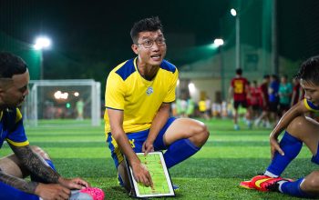 Seorang pelatih sepak bola merumuskan strategi di lapangan bersama pemain selama sesi permainan malam.