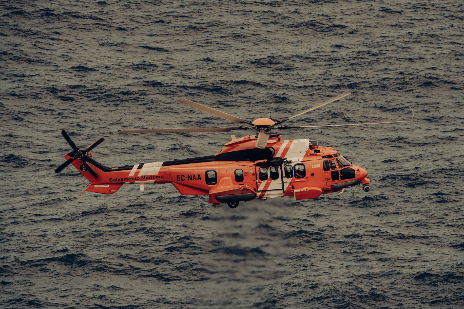 Helikopter penyelamat oranye terbang di atas ombak lautan, melaksanakan operasi darurat.