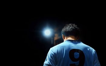 Seorang pemain sepak bola dengan jersey biru bernomor 9, diterangi oleh lampu sorot di malam hari.