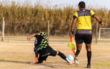 Seorang pemain sepak bola melakukan tekel saat pertandingan, sementara wasit mengamati. Rekaman diambil di luar ruangan, di sebuah lapangan.