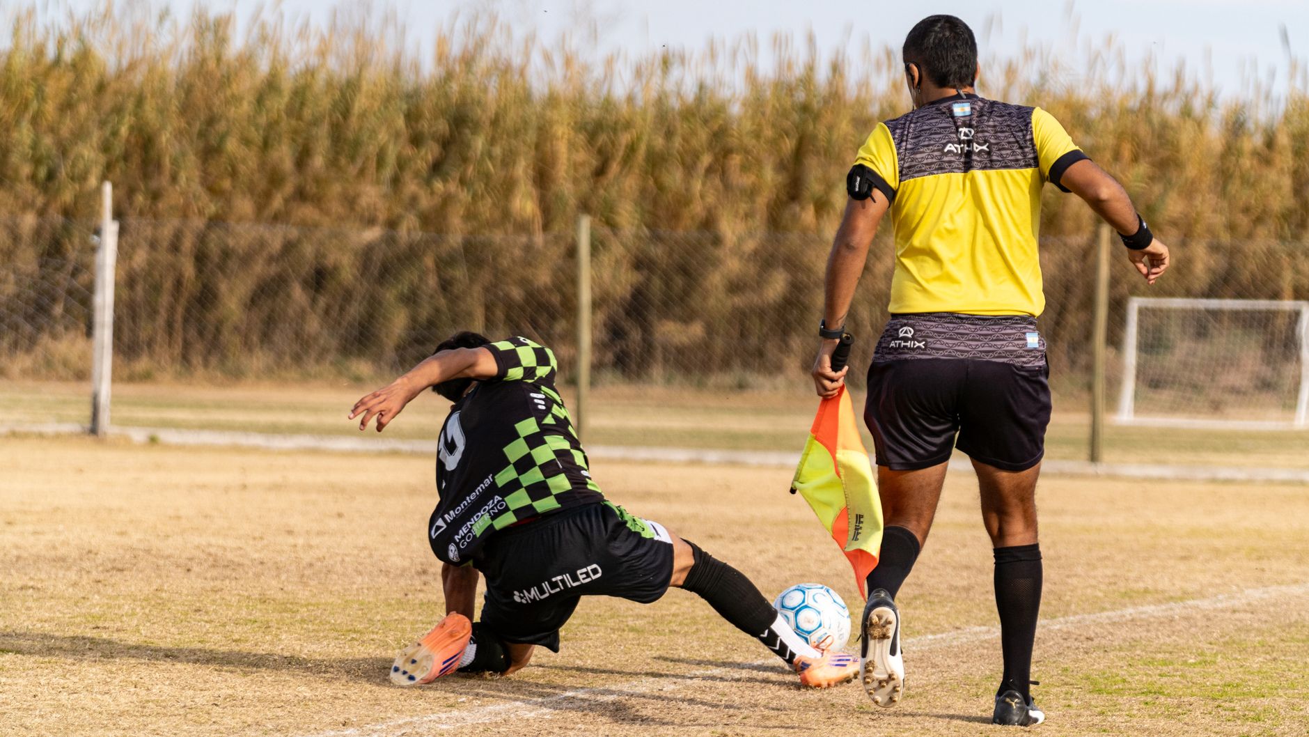 Seorang pemain sepak bola melakukan tekel saat pertandingan, sementara wasit mengamati. Rekaman diambil di luar ruangan, di sebuah lapangan.