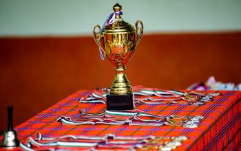 Piala emas dengan pita dan medali di atas meja kotak, melambangkan kemenangan dan pencapaian.