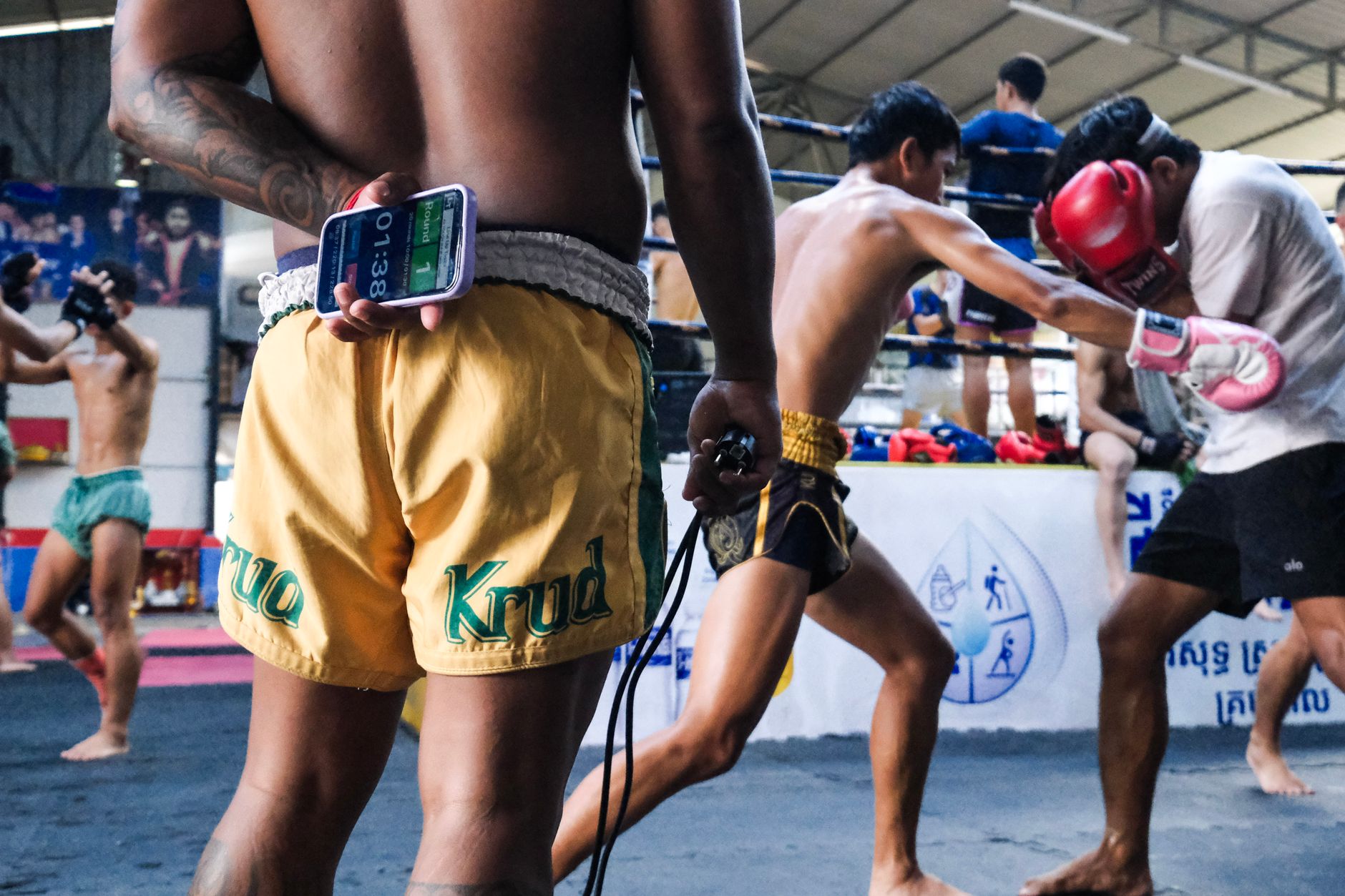 Adegan dinamis para petarung Muay Thai berlatih di sebuah sasana dengan seorang pelatih yang mengatur waktu sesi latihan.