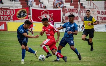 Pemain sepak bola dalam aksi selama pertandingan kompetitif di lapangan rumput dengan penonton di tribun.