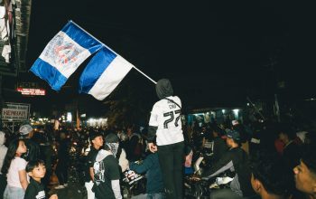 Sebuah kumpulan jalanan yang meriah di malam hari dengan bendera yang ditampilkan secara mencolok di tengah kerumunan besar.