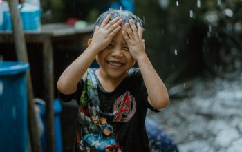 Anak yang tersenyum menikmati hujan dengan pakaian santai, merangkul momen masa kecil yang bebas.
