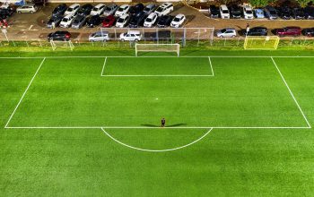 Pemandangan udara lapangan sepak bola di malam hari di Jakarta dengan seorang pemain sendirian yang melemparkan bayangan di bawah lampu stadion.