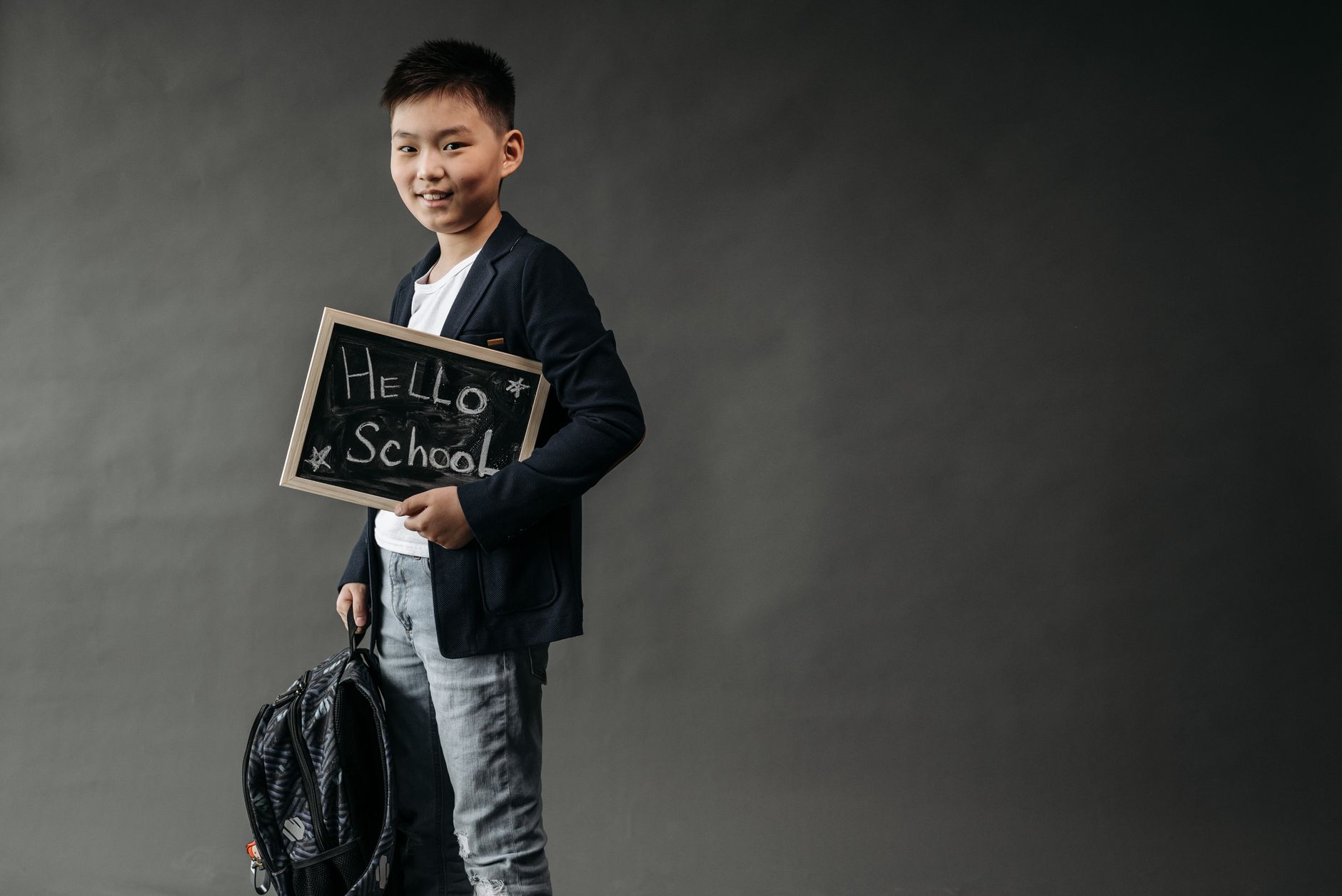 Seorang bocah muda dengan ransel memegang papan tulis bertuliskan 'Halo Sekolah', siap untuk sekolah.