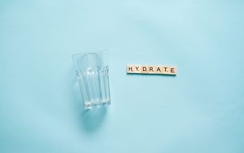 Tata letak minimalis gelas minum dan papan huruf yang mengeja 'HYDRATE' di latar belakang biru.