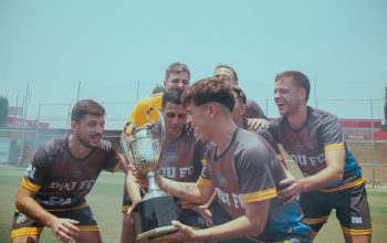 Tim sepak bola dengan gembira merayakan kemenangan mereka di lapangan yang cerah sambil memegang trofi.