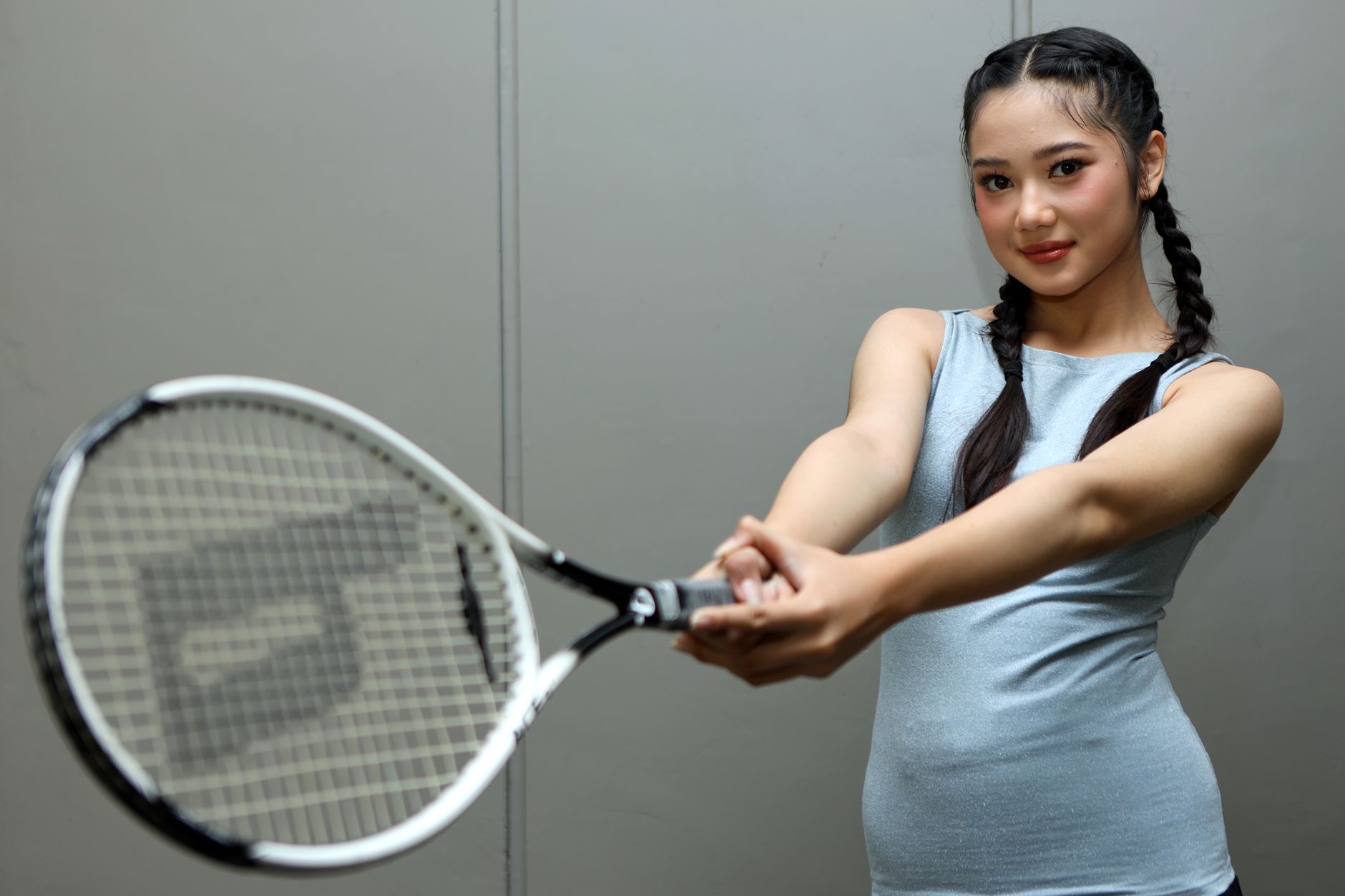 Seorang wanita muda yang percaya diri dengan rambut dikepang memegang raket tenis di dalam ruangan, siap bermain.