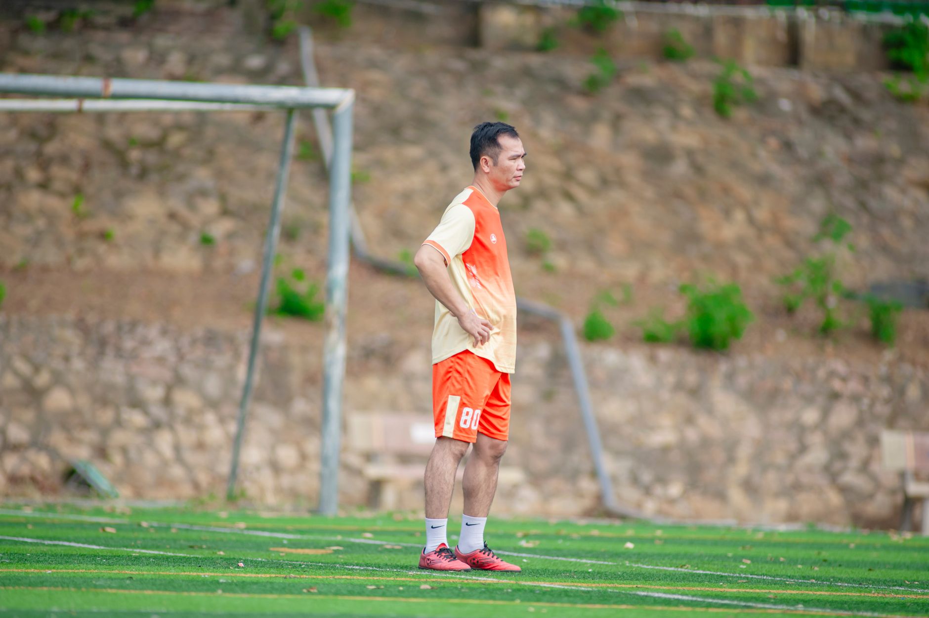 Seorang pria mengenakan pakaian olahraga berdiri di lapangan sepak bola pada siang hari.
