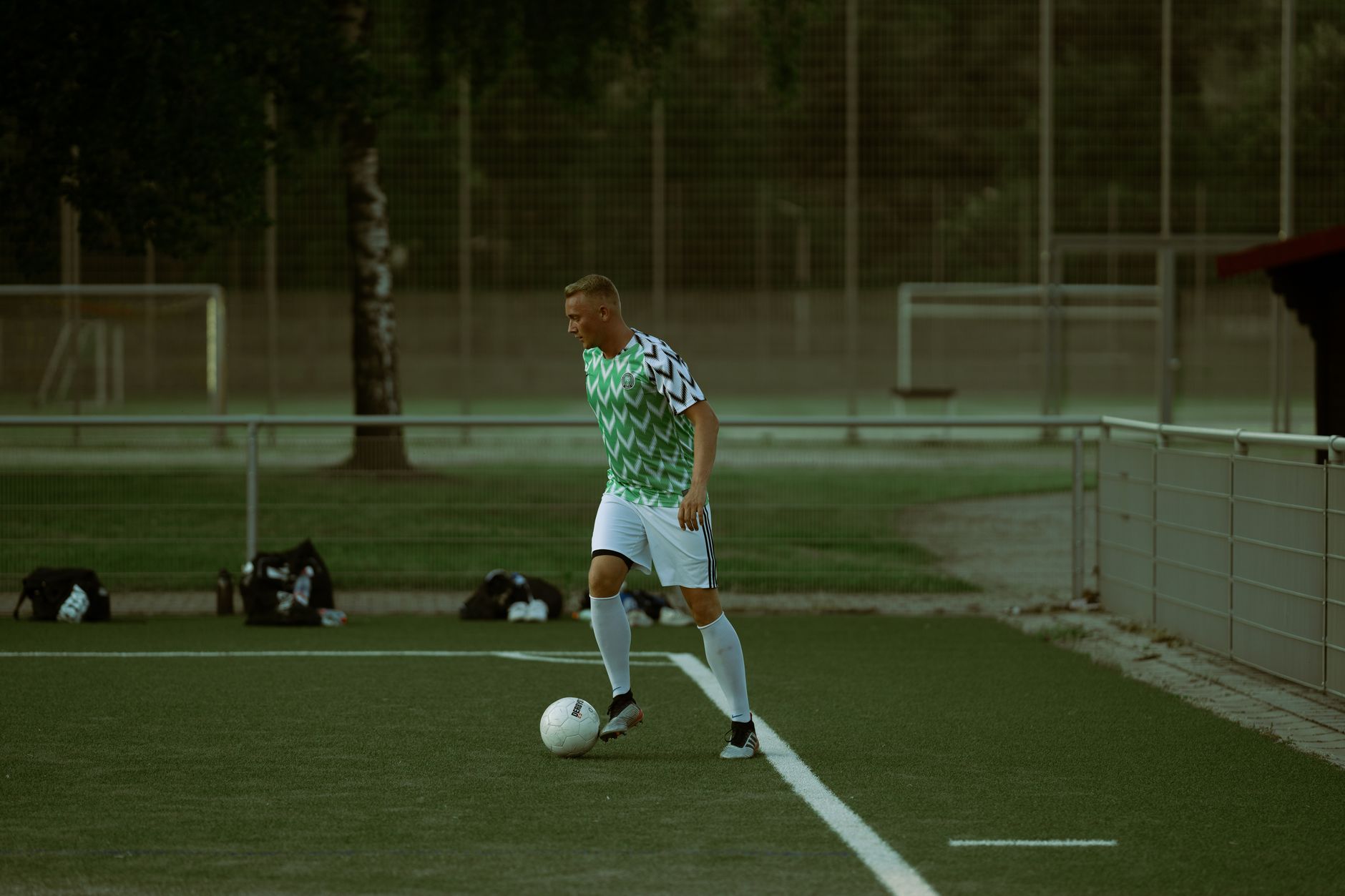 Seorang pemain sepak bola mengiring bola di lapangan hijau pada siang hari, menunjukkan keterampilan atletik.