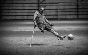 Tangkapan dinamis dari pemain sepak bola adaptif yang menendang bola dengan kruk di dalam ruangan dalam hitam dan putih.