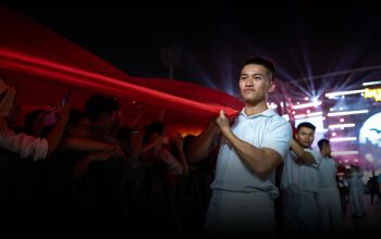 Seorang pemuda gagah berani memegang bendera besar di sebuah acara malam yang meriah dengan lampu-lampu.