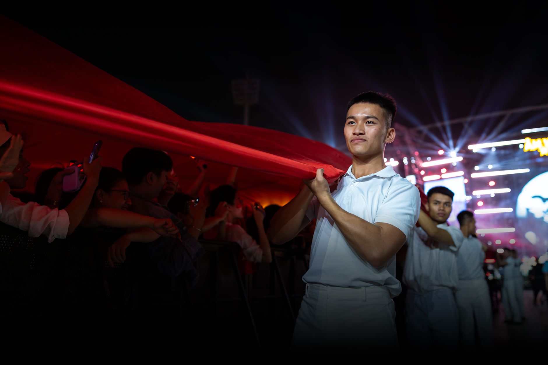 Seorang pemuda gagah berani memegang bendera besar di sebuah acara malam yang meriah dengan lampu-lampu.
