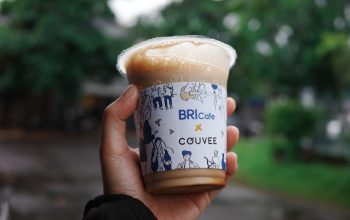 Close-up tangan memegang cangkir kopi es bermerek di luar ruangan, dengan latar belakang hijau buram yang cerah.